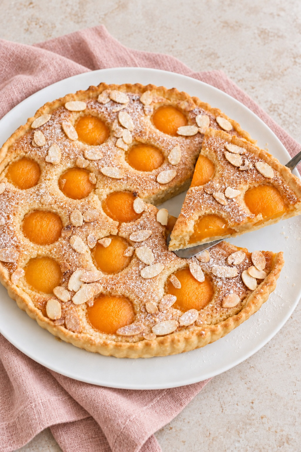 Tarte amandine aux abricots garnie d’amandes effilées et de sucre glace, avec une part découpée et soulevée.
