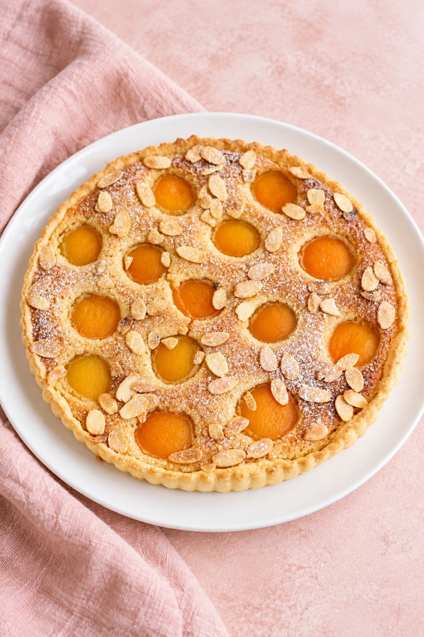 Tarte amandine aux abricots vue du dessus, décorée d’amandes effilées et d’un léger voile de sucre glace.