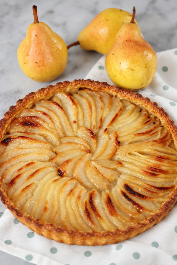 Tarte aux poires fraîches facile, disposées en rosace sur une pâte sablée dorée, avec trois poires en arrière-plan.